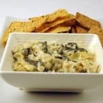 Hot Swiss Chard and Artichoke Dip served with crackers in a white square bowl as appetizers