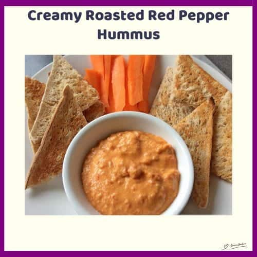 An image of a white bowl filled with Creamy Roasted Red Pepper Hummus with toasted bread triangles and carrots on the side