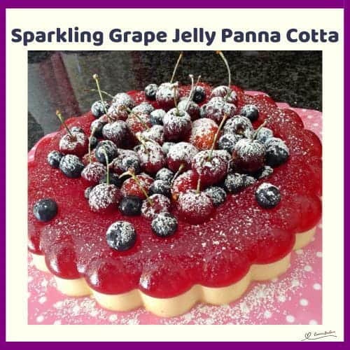 An image of a Sparkling Grape Jelly Panna Cotta, decorated with frosted cherries and fresh berries and a dusting of icing sugar