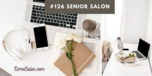 white laptop with black keys on a white desk, with taupe book and white flowers on top of it and a white iphone with black screen and white headphone