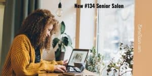 girl with yellow jersey working on laptop sitting on a brown table neat a window with green pot plants to the side