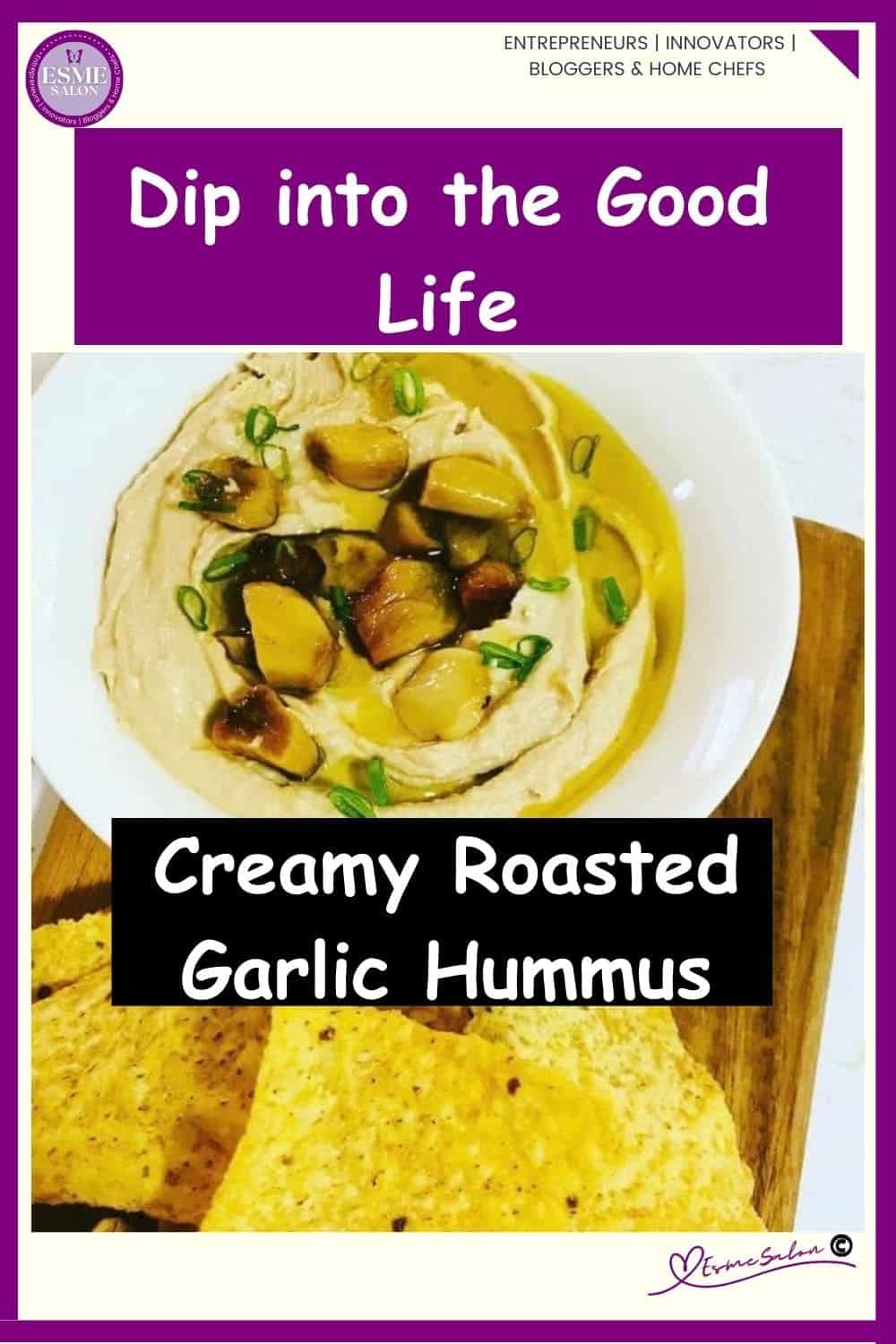 an image of a white bowl filled with Creamy Garlic Roasted Hummus on wooden table with some crisps as well