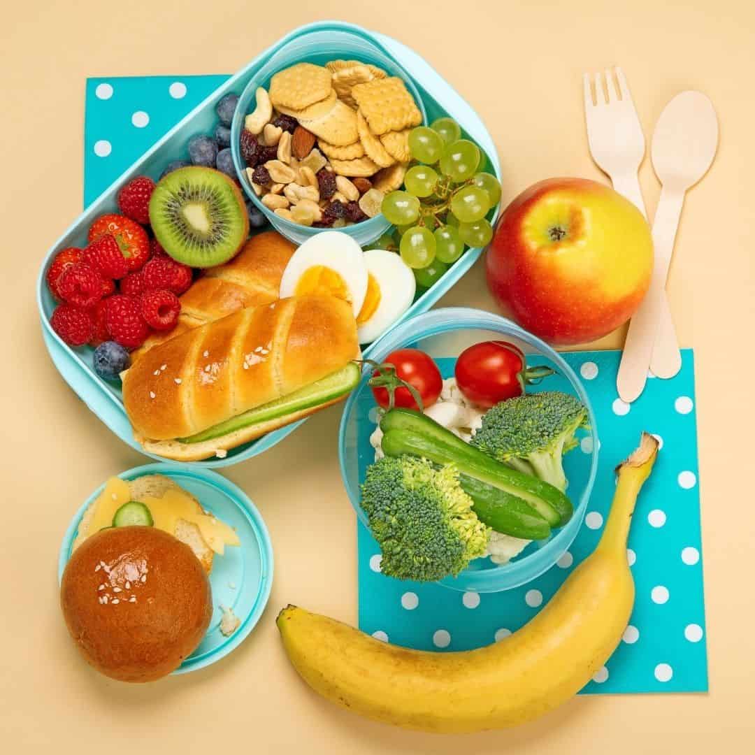 An image with a blue plastic lunch box filled with fruit, crakers, bread, hard boiled eggs with an apple and banana on the side
